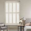 White plantation shutters, four hinged louvered panels covering the window, closed with slats angled to admit diffused light into a neutral bedroom corner with armchair, bedside table, lamp and flowers.