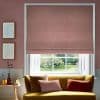 A textured dusty-pink Roman blind is lowered over a white-framed sash window, partially filtering daylight in a living room with a mustard-yellow sofa, cushions, side table and green garden view.