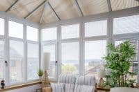 White horizontal blinds cover tall multi‑pane conservatory windows, closed and filtering bright daylight; room contains striped cushioned rattan chairs, wicker lamps, a potted bamboo and suburban roof views.