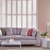 White louvered plantation shutters cover a tall bay window, closed and filtering daylight; in a pastel living room with light sofa, patterned and blush cushions, low coffee table and orchid.
