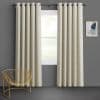 Grommet-top beige curtains, full-length panels on a rod, hung mostly closed with regular folds, filtering soft daylight over a white-framed window showing lawn and trees in a gray-walled room.