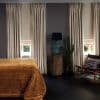 tall beige pleated curtains with lowered Roman shades, partially drawn, softly filtering daylight; bedroom setting with dark floors, rust-colored bedspread, turquoise lamp on a wood stump, potted cactus and chair.
