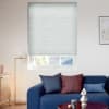 Light-gray pleated cellular blind, lowered almost fully over the window, gently diffusing daylight; in a bright living room with a blue sofa, colored cushions, a floor lamp, and framed art.