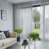 Sheer grey floor-to-ceiling pleated curtains, partly drawn and filtering daylight, revealing a gap to a lawn and shed; modern living room with pale sofa, patterned tile floor and potted plants.