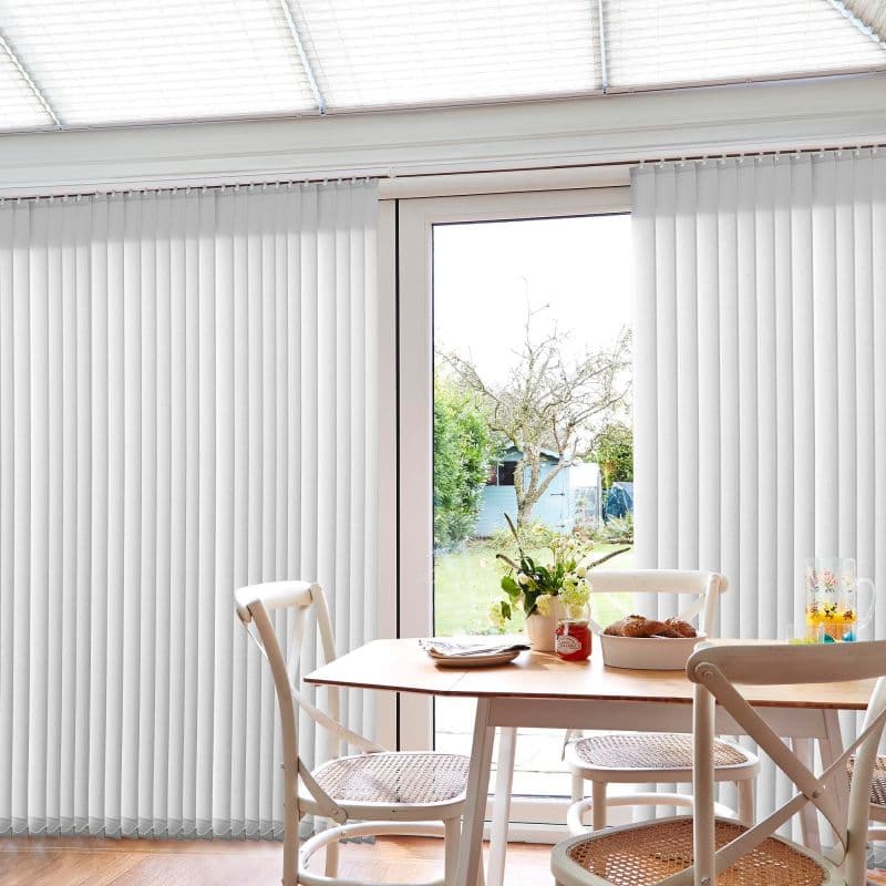 White vertical blinds on a sliding glass door, full-length slatted panels parted at center; filtering bright daylight and pulled aside; dining area with wooden table, chairs, vase and garden view.
