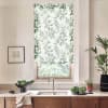 Patterned roller blind with leafy olive motif filters sunlight above a stainless kitchen sink; wooden cabinets, countertop with bowls of greens, a potted plant and framed sketch flank the window.