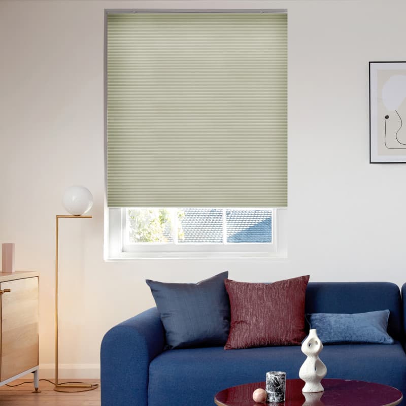 Light-green pleated cellular blind lowered over the window, filtering daylight with a small bottom gap revealing roof and trees; living room with blue sofa, cushions, and a floor lamp.