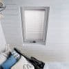 A white pleated skylight blind, fully lowered and slightly diffusing daylight, fitted inside a rectangular roof window; surrounded by white painted wooden tongue-and-groove ceiling above a small bed with cushions.