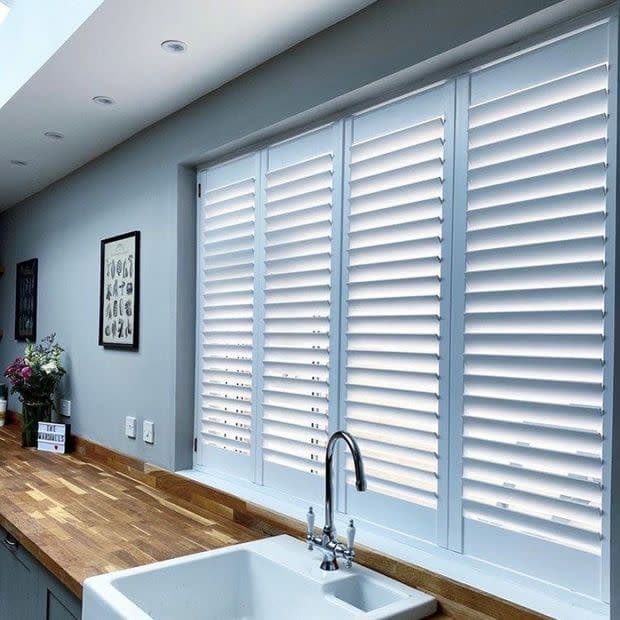 White plantation shutters covering a wide kitchen window, slats tilted to diffuse daylight; farmhouse sink and chrome faucet beneath; wooden countertop, muted grey walls and soft natural light.