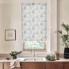 Floral-patterned roller blind covers a window, partially lowered; light filters through foliage outside. Kitchen sink and faucet sit beneath, surrounded by wooden cabinets, countertop plants, bowl of vegetables, framed sketch.