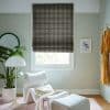 Dark patterned Roman blind, fully lowered with woven geometric motif, shading a white-framed window; pale-green sitting room with white lounge chair, floor lamp, plant and hanging yellow robe, daylight.