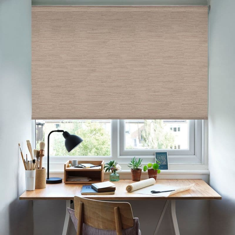 Beige textured roller blind lowered over the window, blocking most light; small home desk beneath with lamp, art supplies, potted plants, notebooks, and a leafy exterior view of nearby buildings.