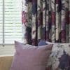 Floral curtains (purple, white peony pattern) hang pleated on the right, partially covering white horizontal blinds filtering daylight; mauve and floral cushions on nearby seating in a softly lit room.