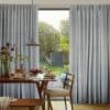 Pleated floor-length grey curtains, drawn open at the center, framing and filtering daylight through a glass door into a dining area with wooden table, pendant lamp, and sunlit garden beyond.