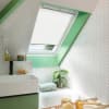 White pleated skylight blind lowered over a slanted roof window, filtering daylight; set in a green-trimmed attic bathroom with white herringbone tiles, sink, bathtub, potted plant, and bath tray.