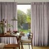 Floor-length pleated mauve curtains, parted to reveal sliding glass door and filter soft daylight. Wooden dining table with pendant lamp, pancakes and vase, opening onto sunlit garden.