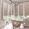 Beige horizontal Venetian blinds covering tall bay windows and doors, mostly lowered and filtering daylight; sunlit dining nook with white table and chairs, floral cushions, tiled floor, garden view.