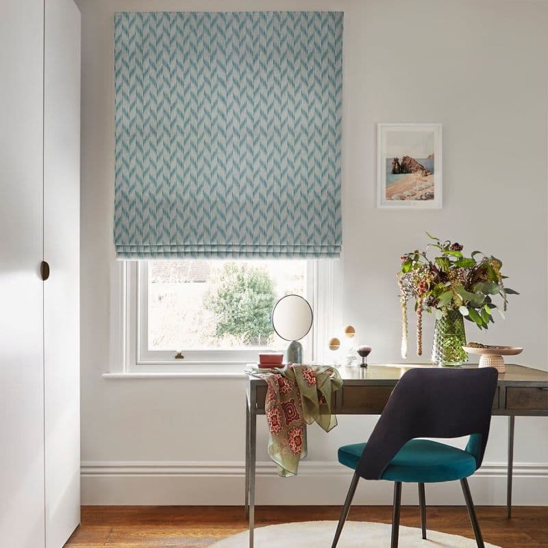 Turquoise patterned Roman blind lowered over a sash window, softly filtering daylight through its lower gap; desk with mirror, vase of flowers and chair define a home office.