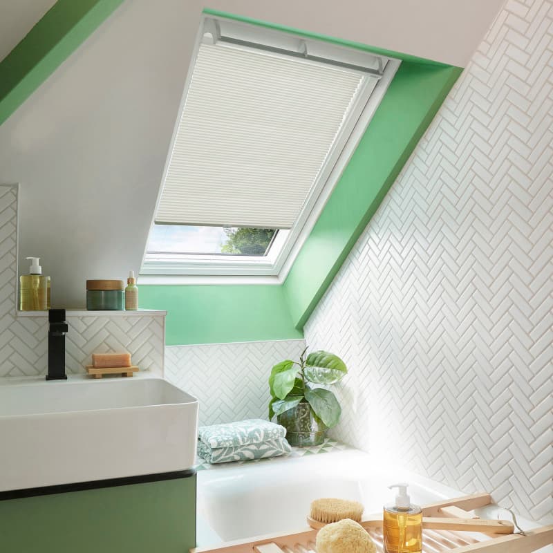 A white pleated skylight blind pulled down halfway, filtering soft daylight; set in a bright attic bathroom with green-painted frame, sloped ceiling, white herringbone tiles, sink, bathtub, and plant.