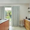 Floor-length patterned white curtains with vertical charcoal brushstrokes, partially drawn across a sliding glass door, letting daylight and a backyard garden view into a bright modern kitchen with wooden cabinets.