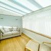Vertical white blinds cover the side window, while pleated translucent roof blinds cross the ceiling, both closed and softly diffusing daylight; a small conservatory with a pale sofa and wooden floor.