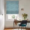 Teal geometric Roman blind, fully lowered, filtering soft daylight through a window; a wooden desk beneath holds a round mirror, vase of flowers, draped scarf, chair in a home office.