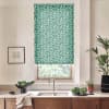 Green floral roller blind covers a kitchen window, filtering daylight; faucet and sink sit beneath on a wooden countertop with potted plants, a bowl of produce and a framed sketch.