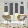 Pleated dark-grey cellular blinds covering tall French doors and upper transom windows, lowered to partially block light, in a bright modern living area with wicker chairs and patterned rug.