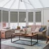 Gray pleated cellular blinds, lowered inside white-framed conservatory windows and filtering sunlight; situated above a beige sofa and wooden coffee table in a softly lit, pastel-accented living room.
