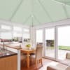 Light-green pleated roof blinds cover the pitched conservatory ceiling, with white pleated blinds on windows and French doors, closed and diffusing daylight over a dining area facing a patio garden.