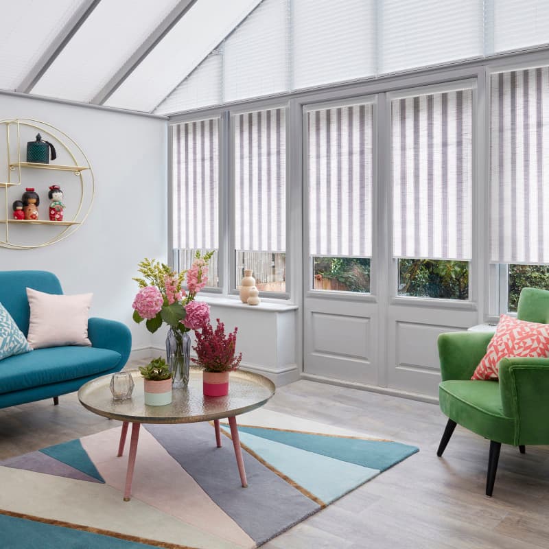 Striped vertical blinds, fully lowered on tall glazed doors and windows, pale-striped; filtering soft daylight in a conservatory with glass roof, teal sofa, green armchair, coffee table and pastel rug.