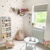 Grey Roman fabric blind (folded tiers) partially lowered, filtering daylight through the window; bright children's nursery with white polka-dot walls, stuffed-animal hammock, bookshelf, beanbag, small table, garden view.
Text found: Maeva
Text found: Dear Zoo