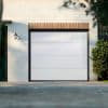 White sectional garage door with dark metal frame closed against a painted white brick façade; trimmed brick header above, wall lantern left, shrub at right, paved driveway and daylight shadows.