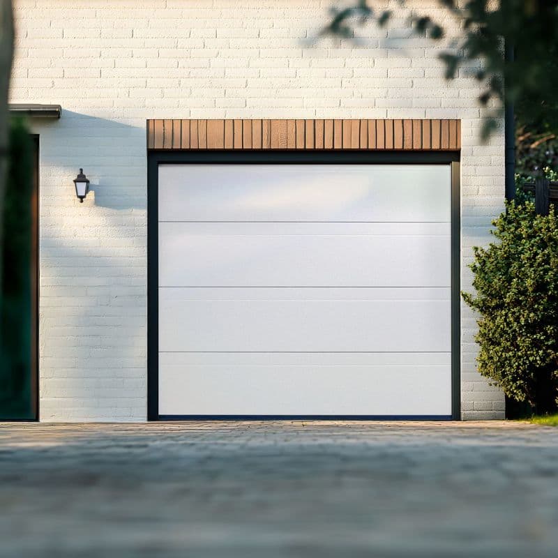White sectional garage door with dark metal frame closed against a painted white brick façade; trimmed brick header above, wall lantern left, shrub at right, paved driveway and daylight shadows.