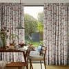 Floral pleated full-length curtains patterned with pink, mauve and grey blossoms frame a sliding glass door, partially drawn to filter light, overlooking a sunlit backyard garden and a breakfast table.