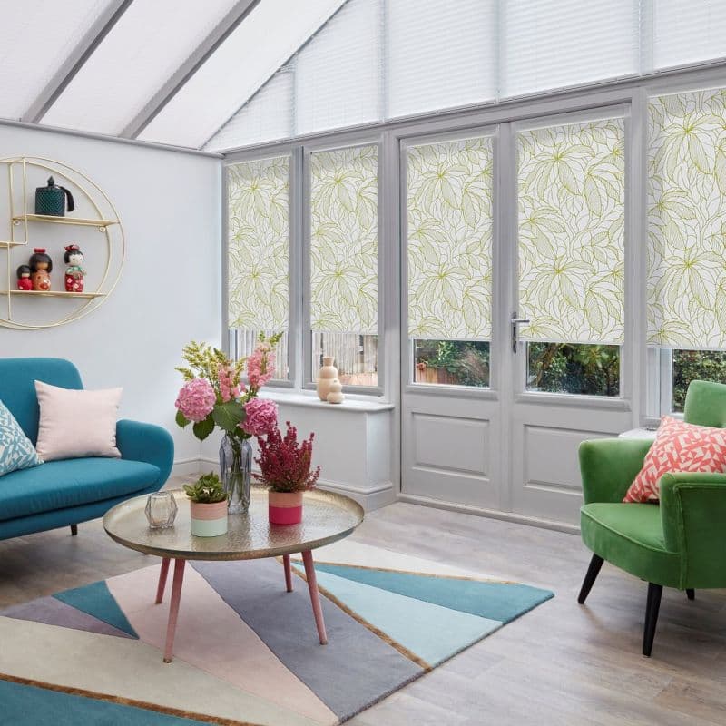 Floral-pattern roller blinds lowered over tall French doors and side windows, with white pleated skylight blinds above; filtering daylight into a bright conservatory-style living room with colorful furniture.
