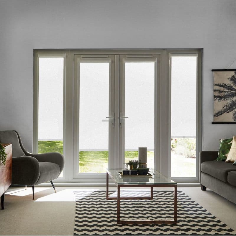White roller blinds on four tall panels (French doors and sidelights), pulled down to diffuse light leaving low clear strips; living room with armchair, glass coffee table and chevron rug.