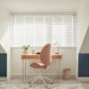 White horizontal blinds across a wide alcove window, closed and filtering bright daylight into striped shadows; small home workspace with wooden desk, pink swivel chair, table lamp, potted plant, walls.