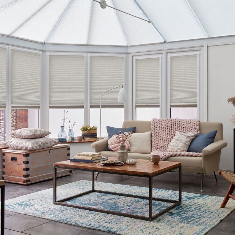 Pleated cellular blinds across conservatory roof and tall bay windows — lowered, filtering soft daylight — in a sunlit living room with beige sofa, chunky pink throw, coffee table on rug.