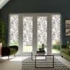 Patterned floral roller blinds cover four tall patio-door panels, filtering daylight in a modern living room with olive-green armchairs, a glass coffee table, zigzag rug, and a garden beyond.