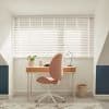 White horizontal blinds span the window, closed with thin slats and pull cord, filtering daylight and casting striped shadows over a wooden desk and pink swivel chair in an attic study.