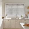 White horizontal Venetian blinds fully covering a large window, slats slightly tilted to filter soft daylight; above a cream kitchen sink and marble countertop with faucet, plants, and utensils.