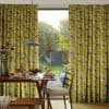 Floral pleated full-length curtains (mustard yellow with red and blue flowers) pulled aside, framing a glass sliding door; dining table with breakfast setting under a pendant lamp, sunlit garden beyond.