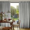 Floor-length grey pleated curtains, drawn slightly apart and gently filtering light; framing a sunlit dining room with wooden table set for breakfast and a view of a lush backyard garden.