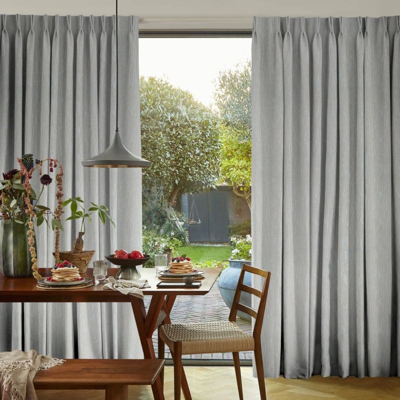 Floor-length grey pleated curtains, drawn slightly apart and gently filtering light; framing a sunlit dining room with wooden table set for breakfast and a view of a lush backyard garden.