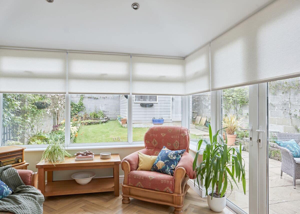 Cream roller blinds cover the upper halves of corner conservatory windows, lowered and softly filtering daylight; inside, an upholstered armchair, wooden table, potted plants, and a small garden beyond.