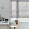 Sheer grey Roman blind, lowered with horizontal ribs, filtering soft daylight and faint tree silhouettes; in a minimalist home office with white desk, wooden chair and a paper pendant.