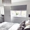 Grey Roman blind, lowered over a three-panel window with a fold at the bottom; filtering daylight into a neutral bedroom, bed and cushions, white vase of flowers on the sill, suburban view.