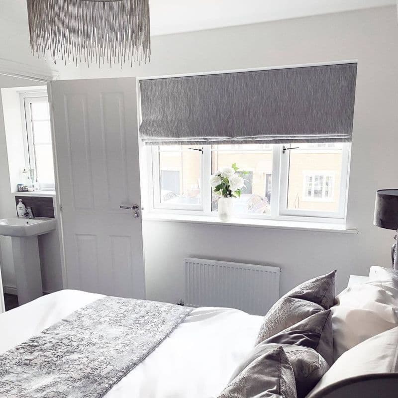 Grey Roman blind, lowered over a three-panel window with a fold at the bottom; filtering daylight into a neutral bedroom, bed and cushions, white vase of flowers on the sill, suburban view.