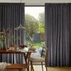 Charcoal-gray pleated curtains with textured weave, drawn partly open framing a glass door; filtering soft daylight into a dining room with a wooden table and leafy garden view.